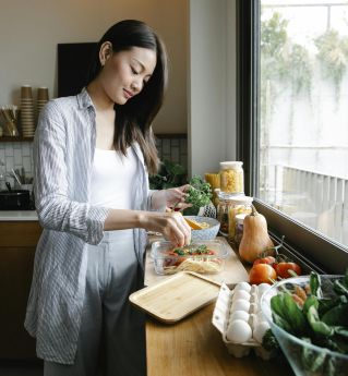 Side view of Asian female adding fresh parsley into glass container with pasta and bolognese sauce while cooking in kitchen with various products and kitchenware